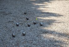 Metallene Boulekugeln und gelbe Zielkugel auf einem Kiesplatz – Symbolbild