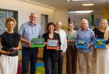Sieben Personen halten Tablets mit Kommunikations-Apps in einer Kita-Garderobe