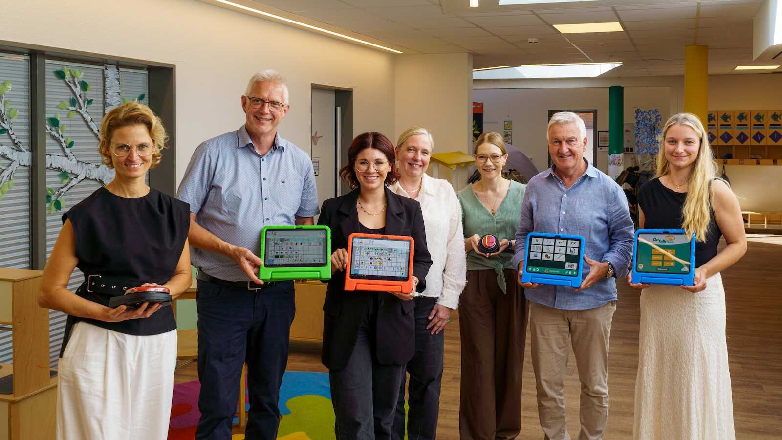 Sieben Personen halten Tablets mit Kommunikations-Apps in einer Kita-Garderobe