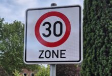 Verkehrsschild Tempo-30-Zone am Straßenrand vor Bäumen und Wohnhäusern
