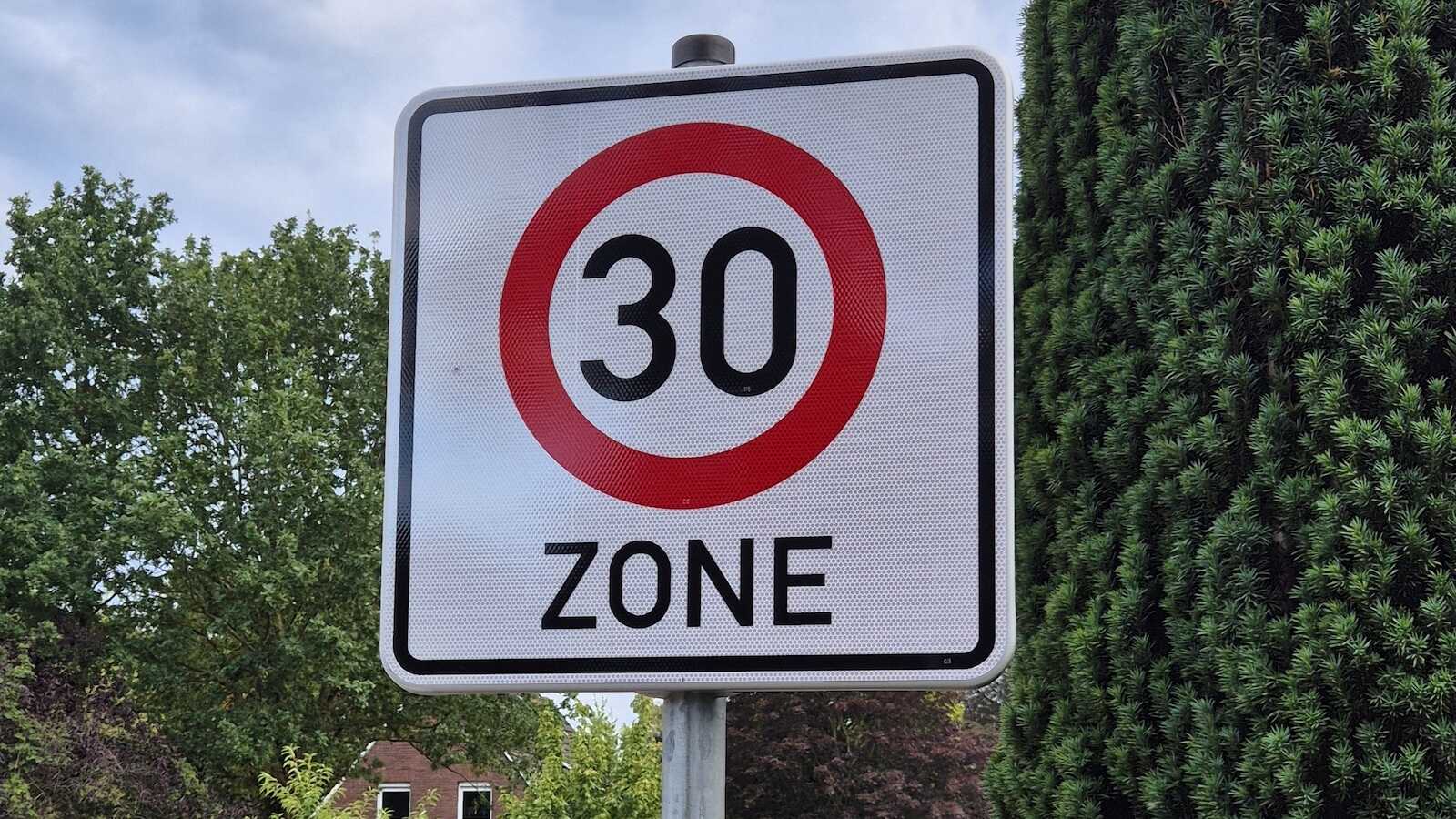 Verkehrsschild Tempo-30-Zone am Straßenrand vor Bäumen und Wohnhäusern