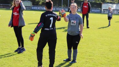 Mannschaften und Zuschauer stehen sich auf dem Sportplatz gegenüber, Spieler heben die Hand