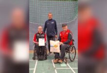 Thomas Hilmes mit Urkunde und Geschenktasche im Rollstuhl, daneben Willi Fenslage und Günter Hömmeke in der Sporthalle