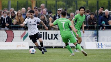 Anton Popov vom SC Spelle-Venhaus im weißen Trikot am Ball, zwei Gegenspieler in Grün laufen auf ihn zu