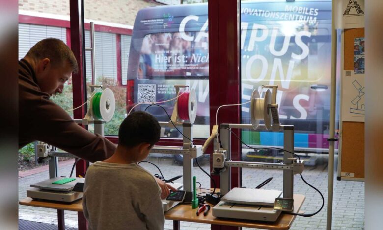 Ein Junge arbeitet mit einem Betreuer an 3D-Druckern, im Hintergrund der Transporter Campus on Wheels