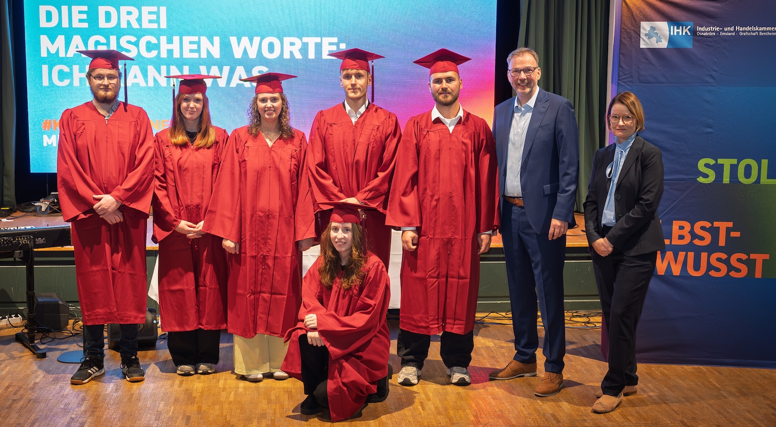 Berufsbeste aus der Grafschaft Bentheim in roten Talaren vor der IHK-Leinwand, rechts IHK-Vizepräsident und Geschäftsbereichsleiterin