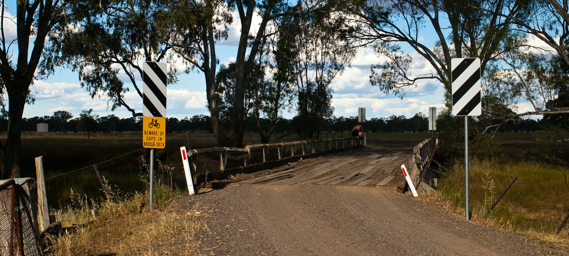 Schmale Brücke auf einem unbefestigten Weg mit Warnschildern, umgeben von Bäumen