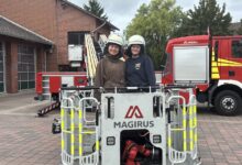 Zwei junge Frauen mit Feuerwehrhelmen stehen hinter dem Rettungskorb einer Drehleiter vor der Feuerwache Lingen