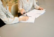 Eine Person unterschreibt ein Dokument, eine zweite Person hält die Unterlagen am Tisch
