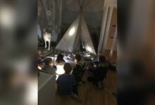 Kinder sitzen im dunklen Museum vor einem Zelt und einem Rentier und leuchten mit Taschenlampen
