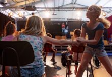 Kinder proben eine Theaterszene mit Stühlen auf einer Bühne mit Scheinwerfern
