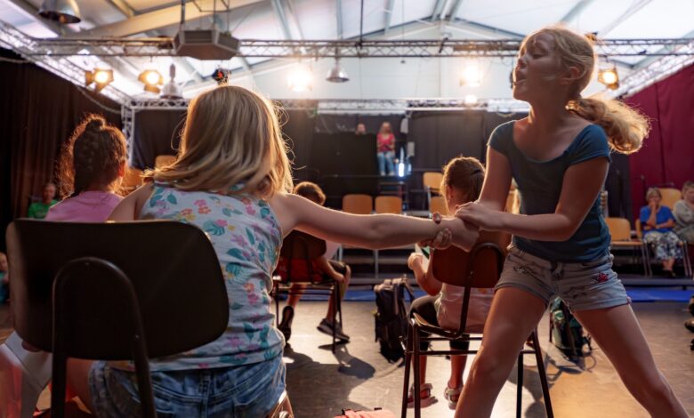 Kinder proben eine Theaterszene mit Stühlen auf einer Bühne mit Scheinwerfern
